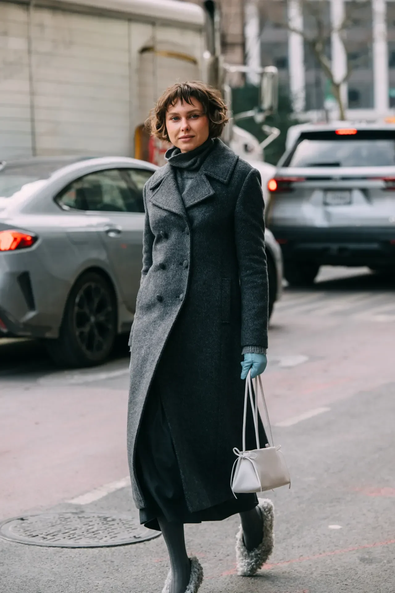 Mulher caminha na rua com casaco cinza longo, luvas azuis de couro, bolsa branca e sapatos peludos, em clima urbano frio.