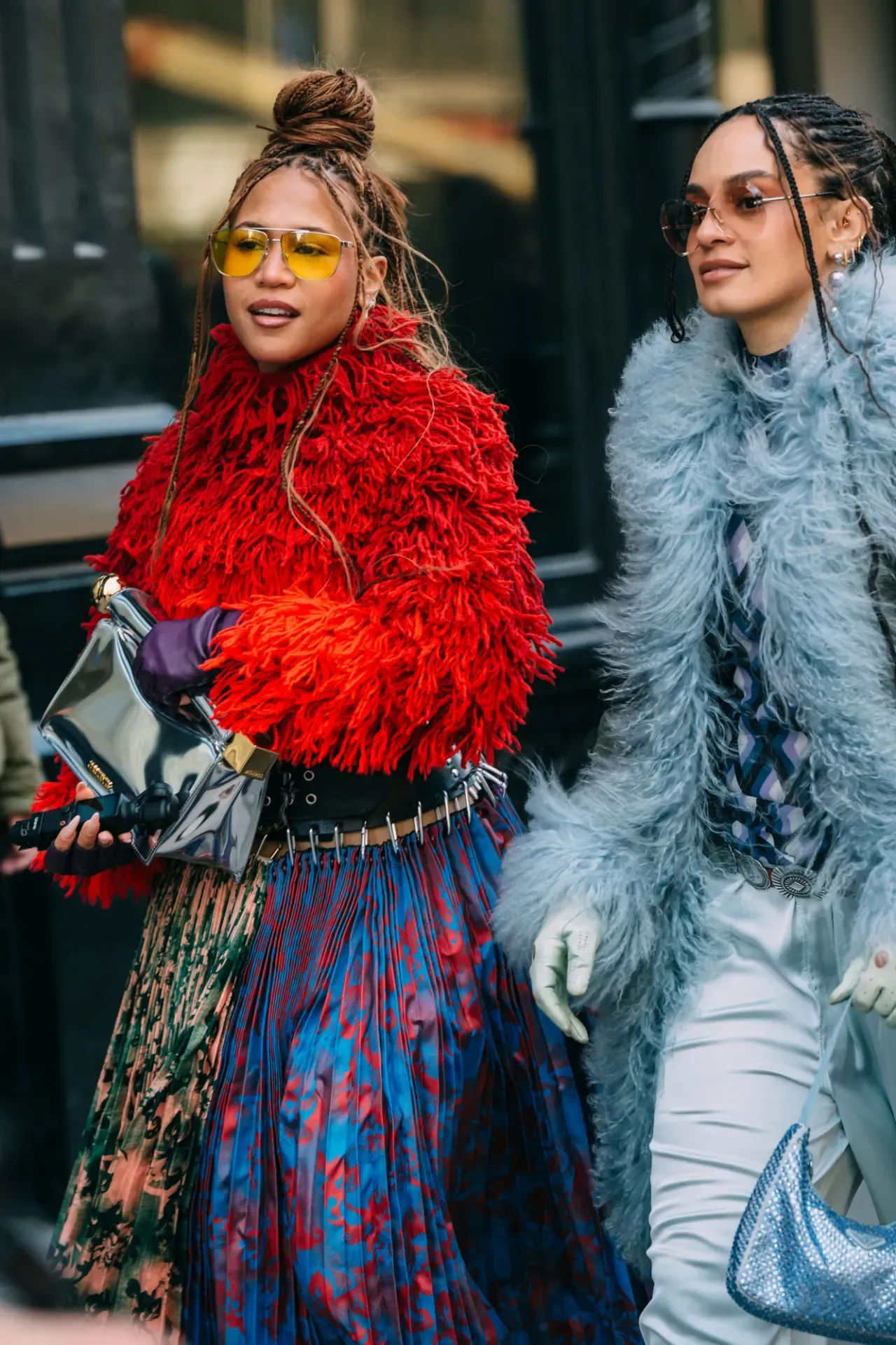 Duas mulheres caminham com estilo usando luvas de couro roxo e branco, casacos peludos vermelho e azul claro e saias e