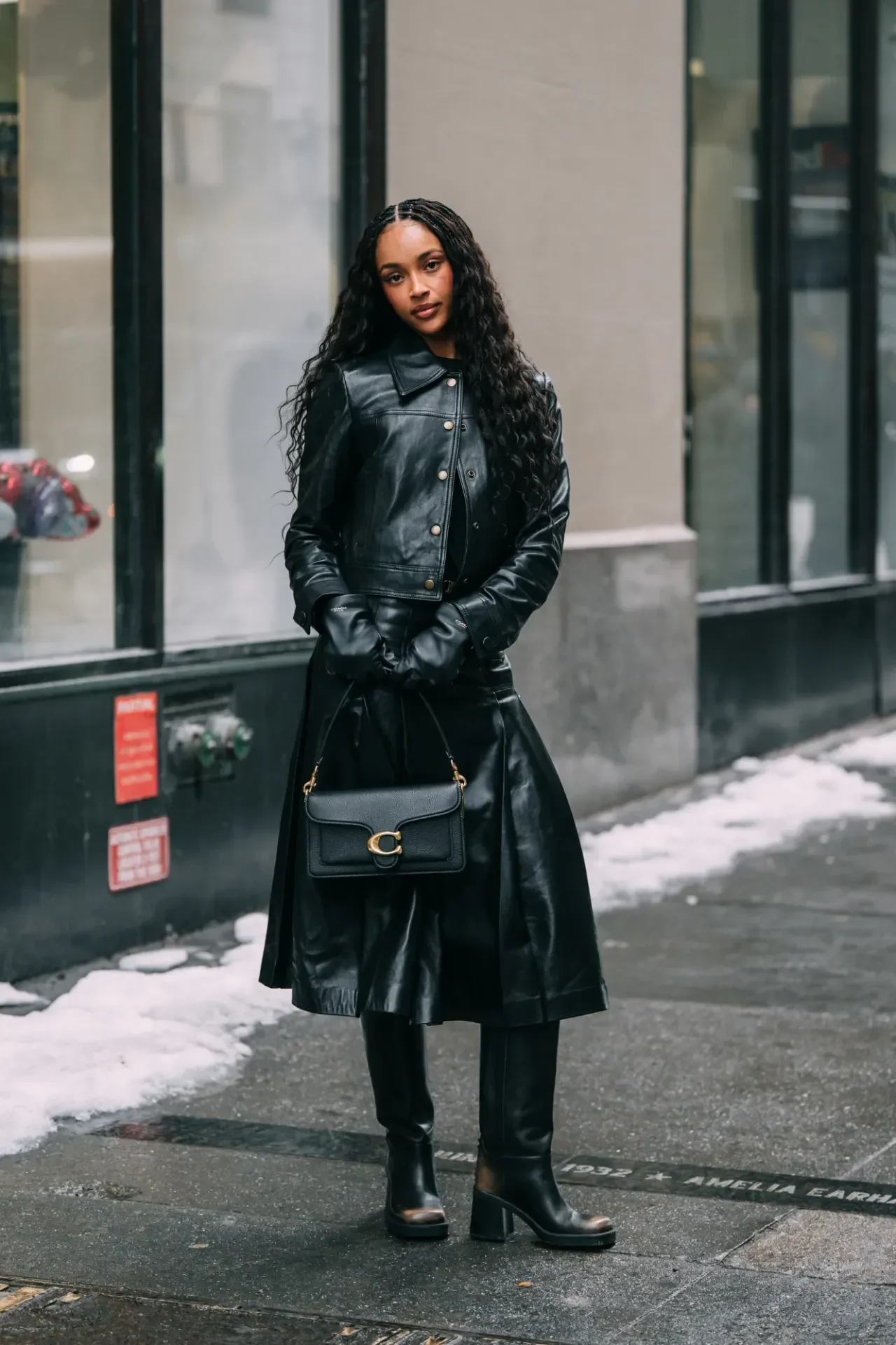 Modelo posa com luvas de couro pretas, jaqueta cropped e saia midi em couro, combinando com botas e bolsa, em clima urbano