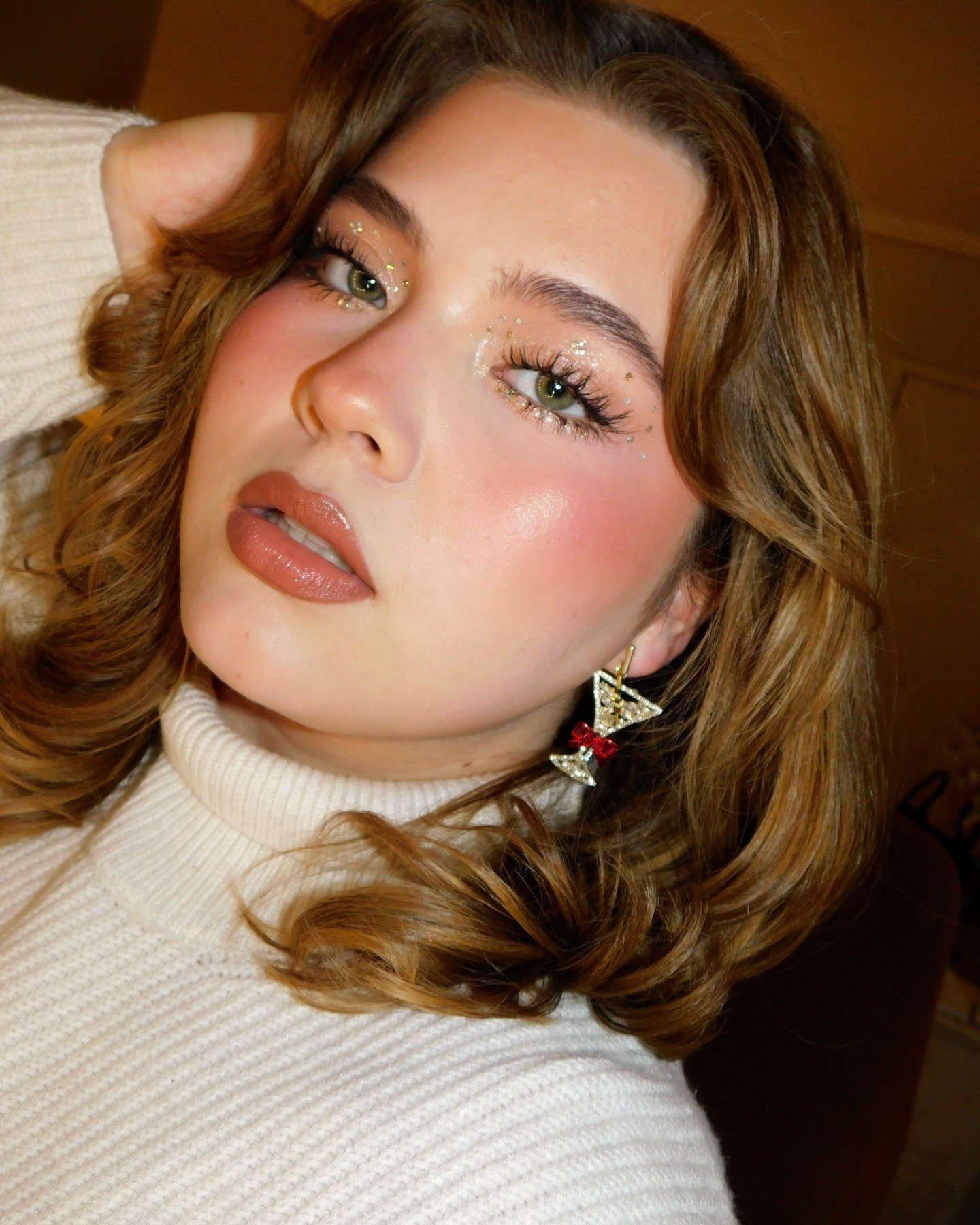 Modelo posa com make de carnaval suave em tons terrosos, glitter dourado nos olhos e blush rosado, combinando com suéter