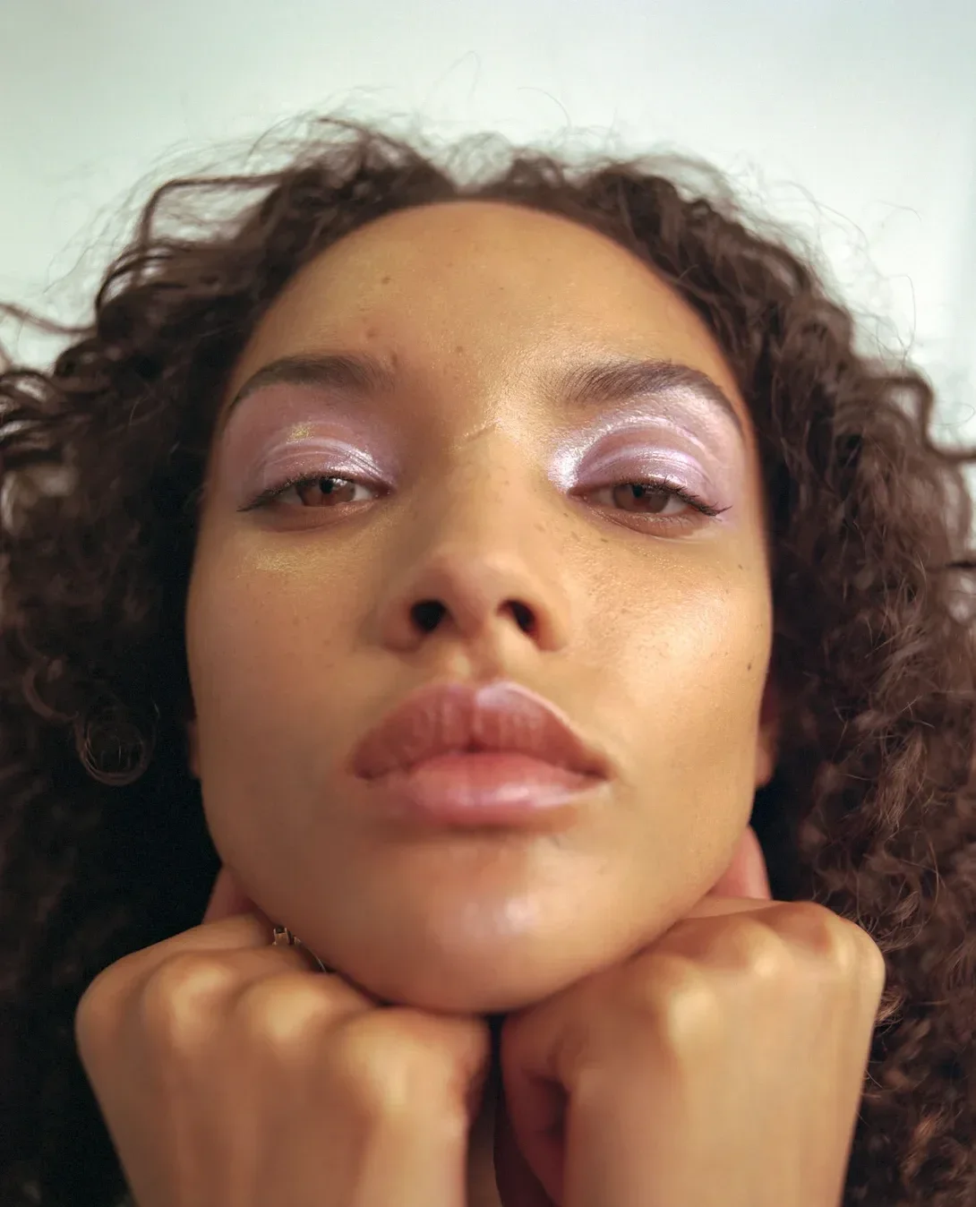 Modelo com make de carnaval em tons perolados lilás nos olhos, pele natural e expressão serena, com mãos apoiando o queixo.