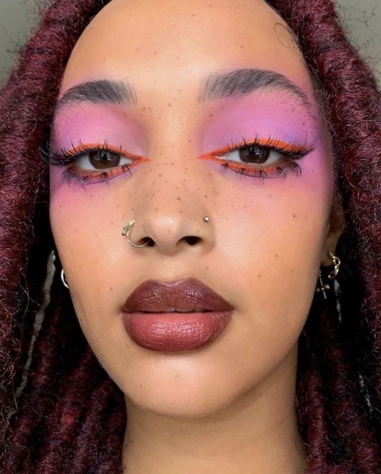 Make de carnaval com sombra rosa vibrante e delineado laranja, lábios marrons e pele com sardas naturais