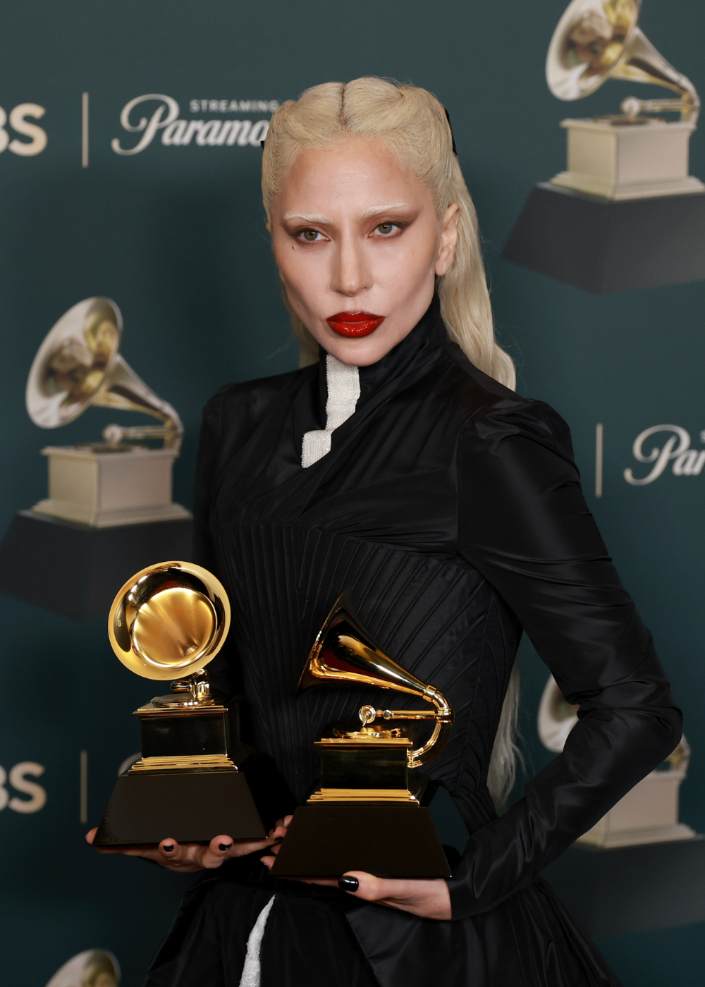 Modelo posa com maquiagem gótica marcada por batom vermelho vibrante, pele pálida, sobrancelhas claras e olhos esfumados.