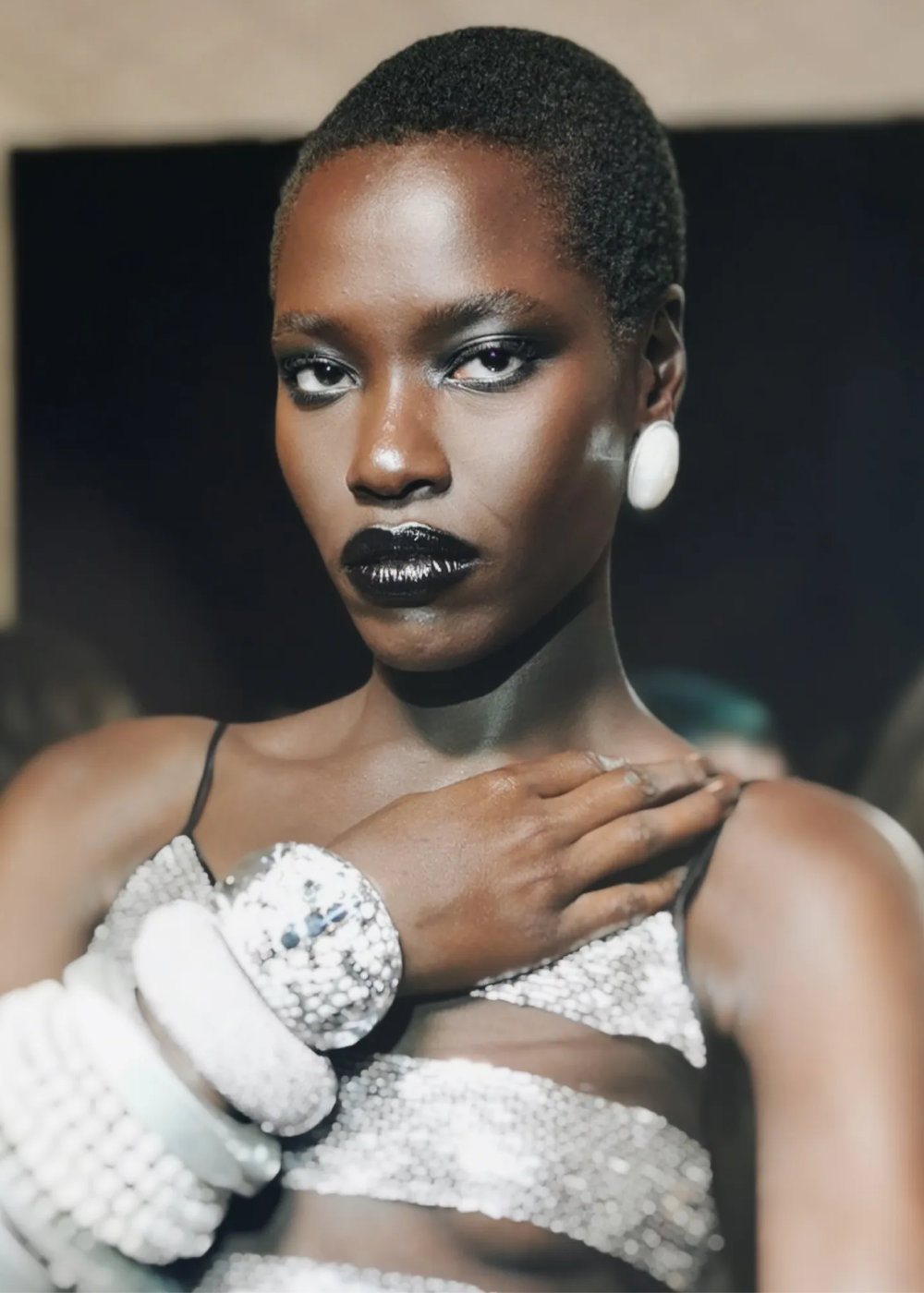 Modelo posa com maquiagem gótica marcada por batom preto brilhante e sombra esfumada, usando top e pulseiras prateadas.