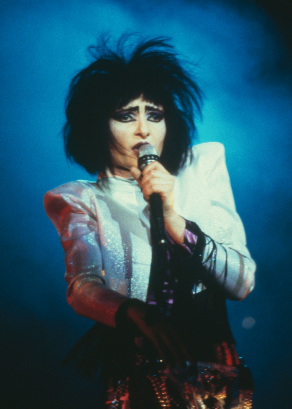 Modelo posa cantando com maquiagem gótica marcada por olhos dramáticos em preto, sobrancelhas fortes e cabelos volumosos.