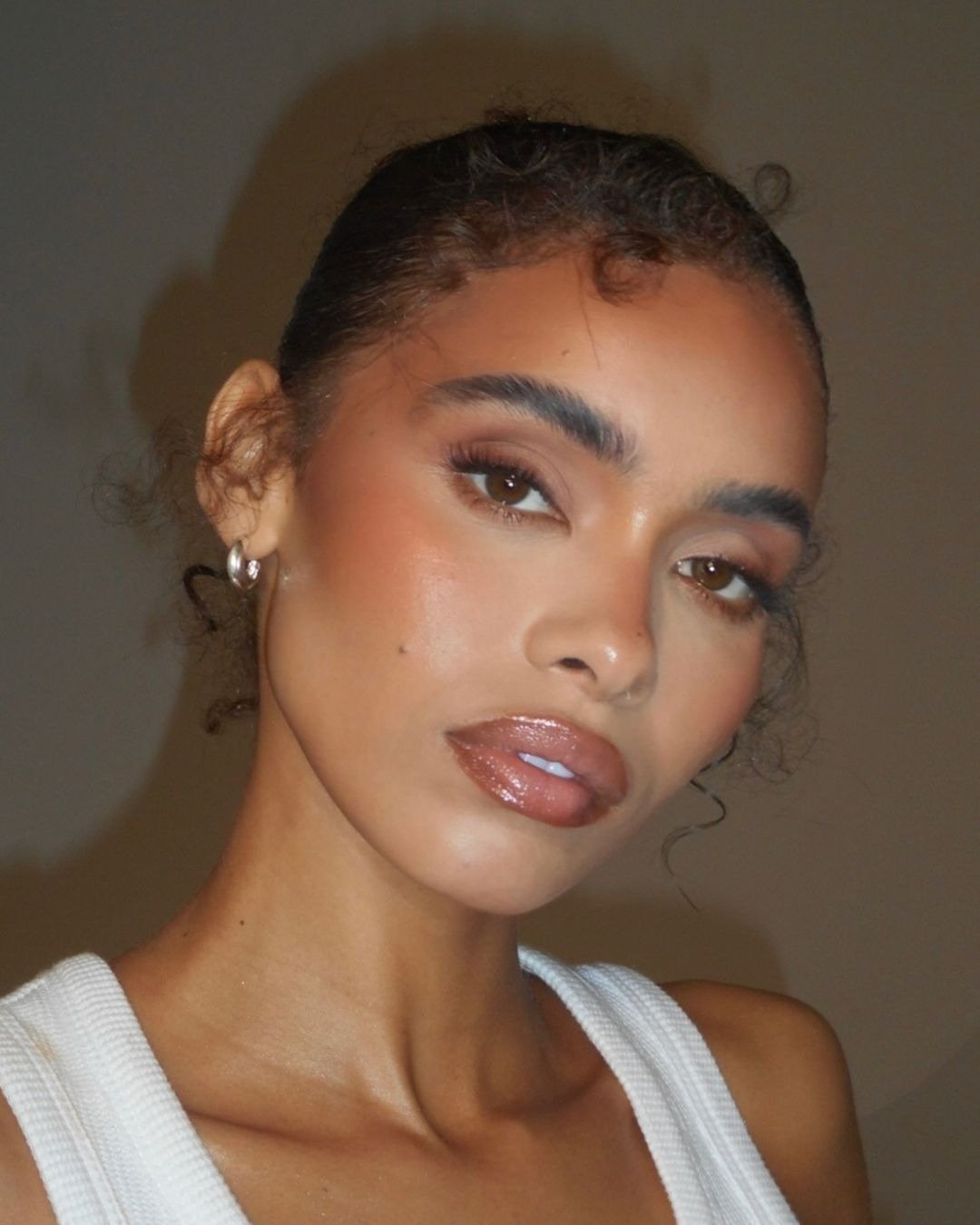 Modelo posa com maquiagem simples em tons terrosos, pele iluminada, batom gloss marrom e regata branca de malha