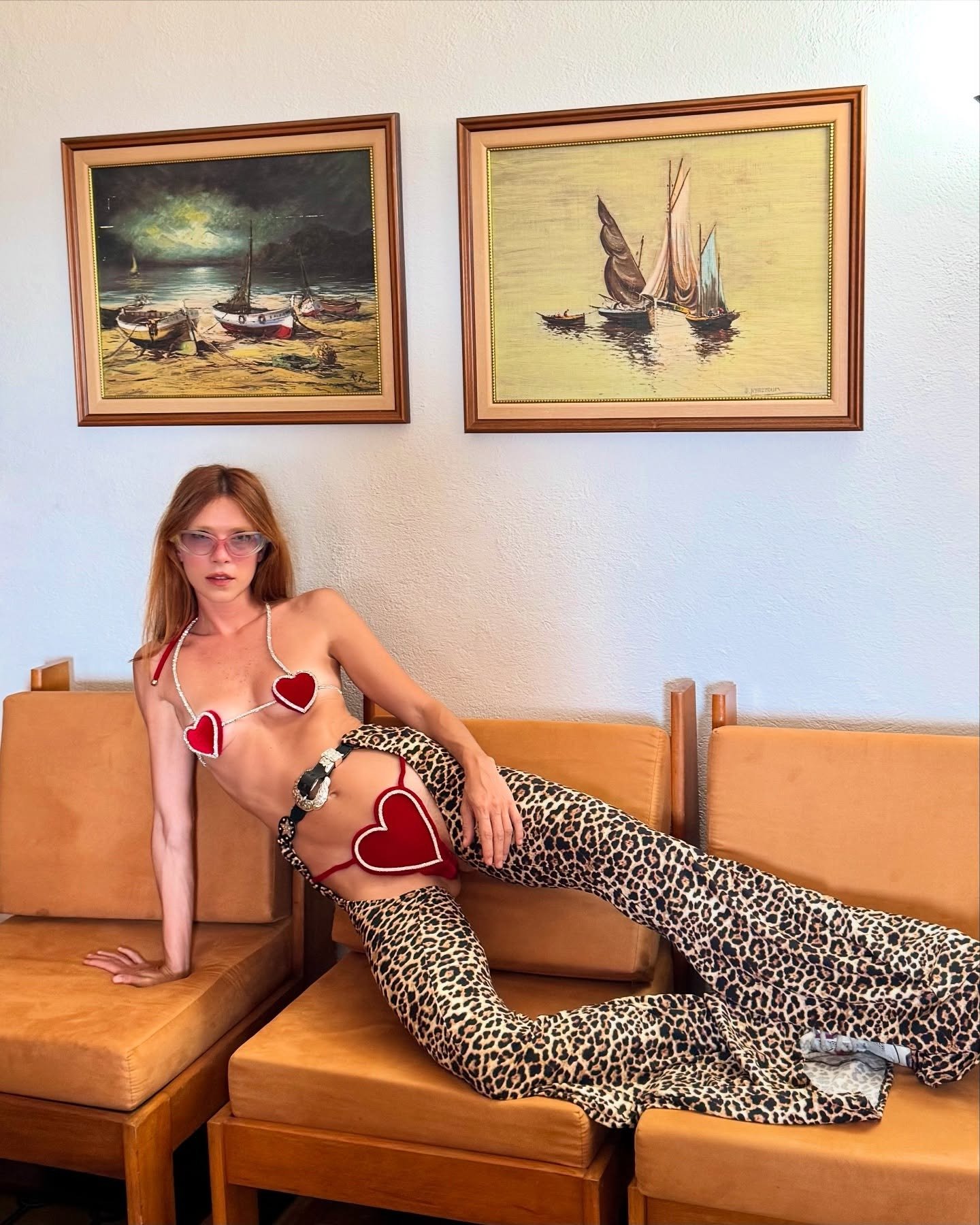 Modelo posa reclinada em sofá caramelo com top de corações vermelhos e calça flare animal print, combinando sensualidade e