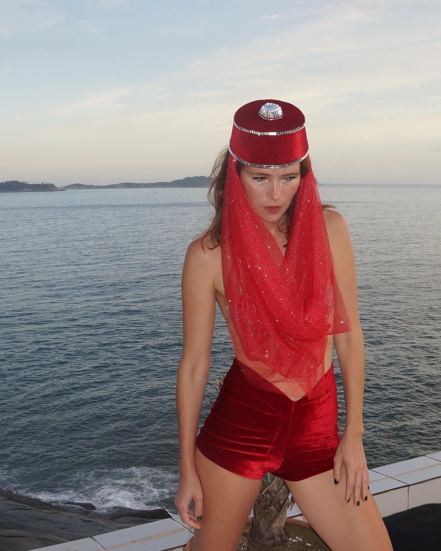 Modelo posa à beira-mar com shorts de veludo vermelho e chapéu com véu de tule brilhante, combinando estilo ousado e festivo