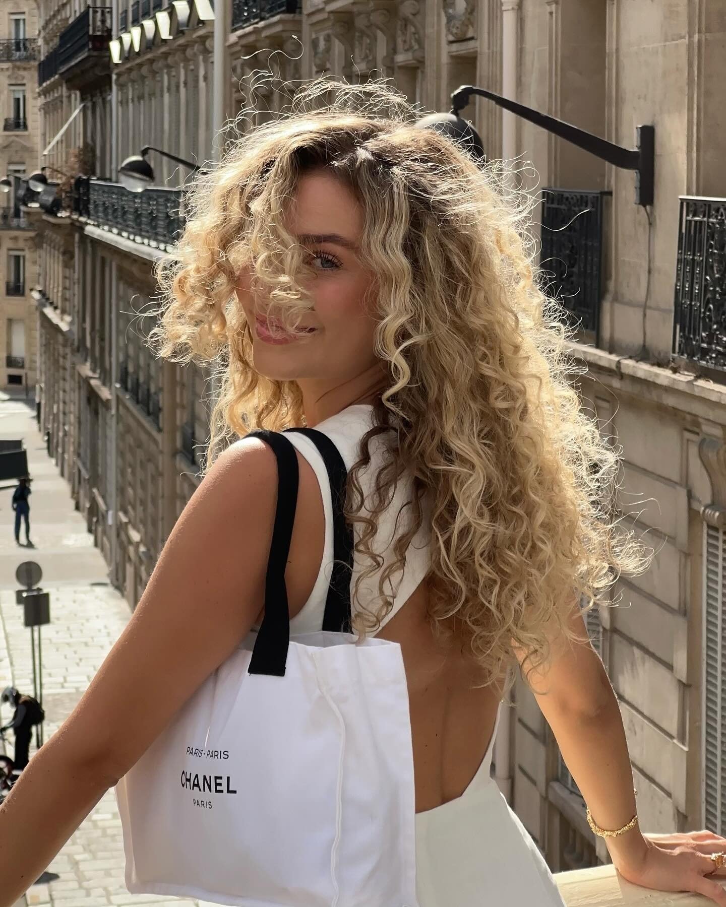 Modelo posa sorrindo com vestido branco de costas nuas e bolsa Chanel branca, cabelo loiro tratado com máscara matizadora