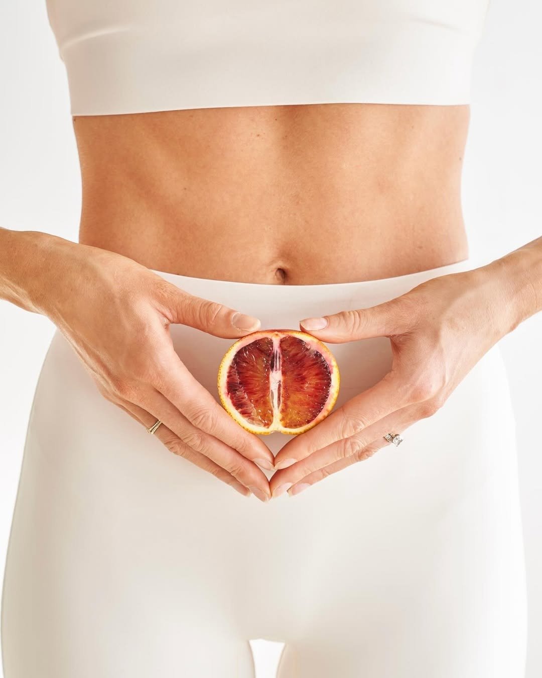 Corpo feminino veste top e legging branca em tecido liso, mãos seguram meio fruto vermelho simbolizando menopausa precoce