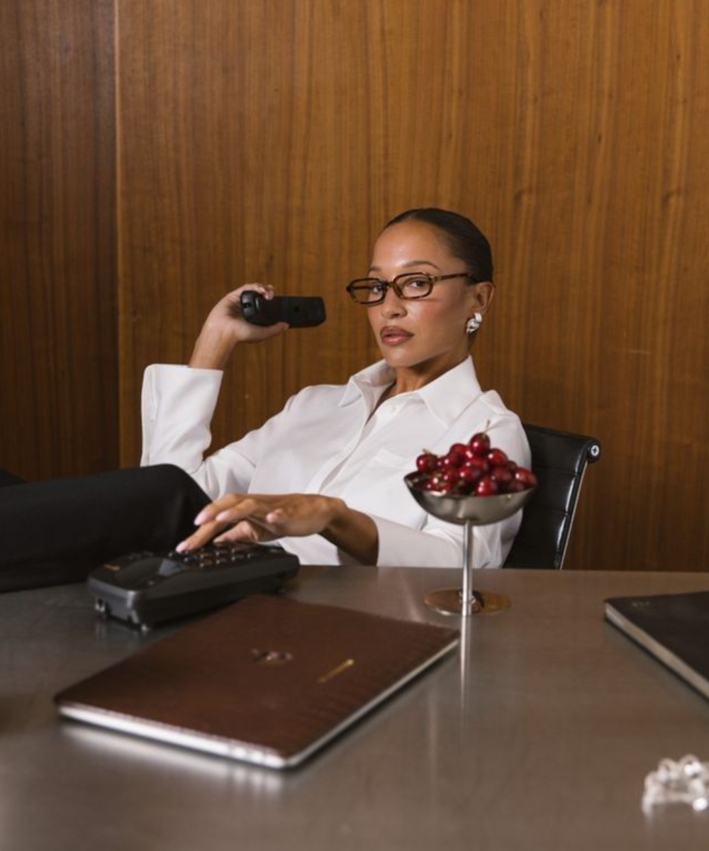 Mulher com camisa branca de modelagem clássica, óculos de armação escura e expressão confiante.