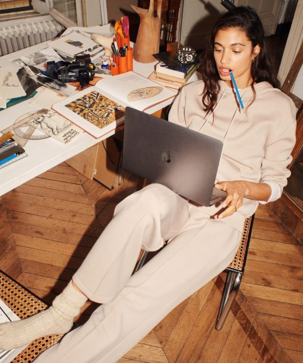 Mulher veste conjunto bege de moletom e meias felpudas, sentada relaxada com laptop, explorando o mercado preditivo na moda.