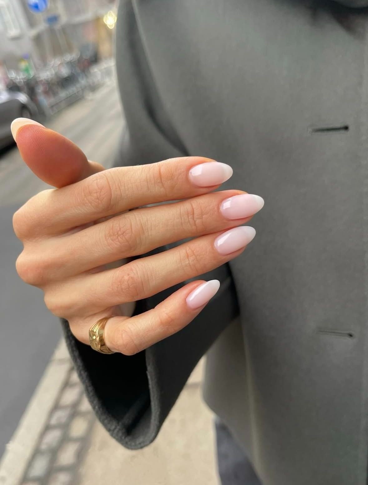 Mão com milky nails em formato amendoado, esmalte translúcido rosado, combinando com casaco cinza e anel dourado, estilo