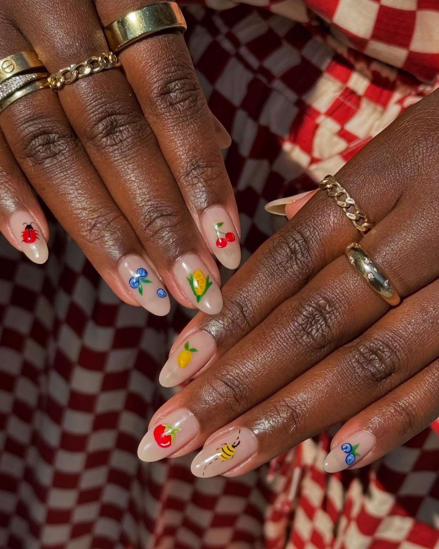 Unhas milky nails em formato amendoado exibem desenhos delicados de frutas e insetos coloridos, com anéis dourados e fundo
