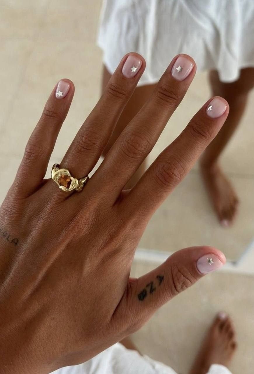 Unhas milky nails curtas com esmalte translúcido e estrelas prateadas, mão adornada por anel dourado em fundo neutro.