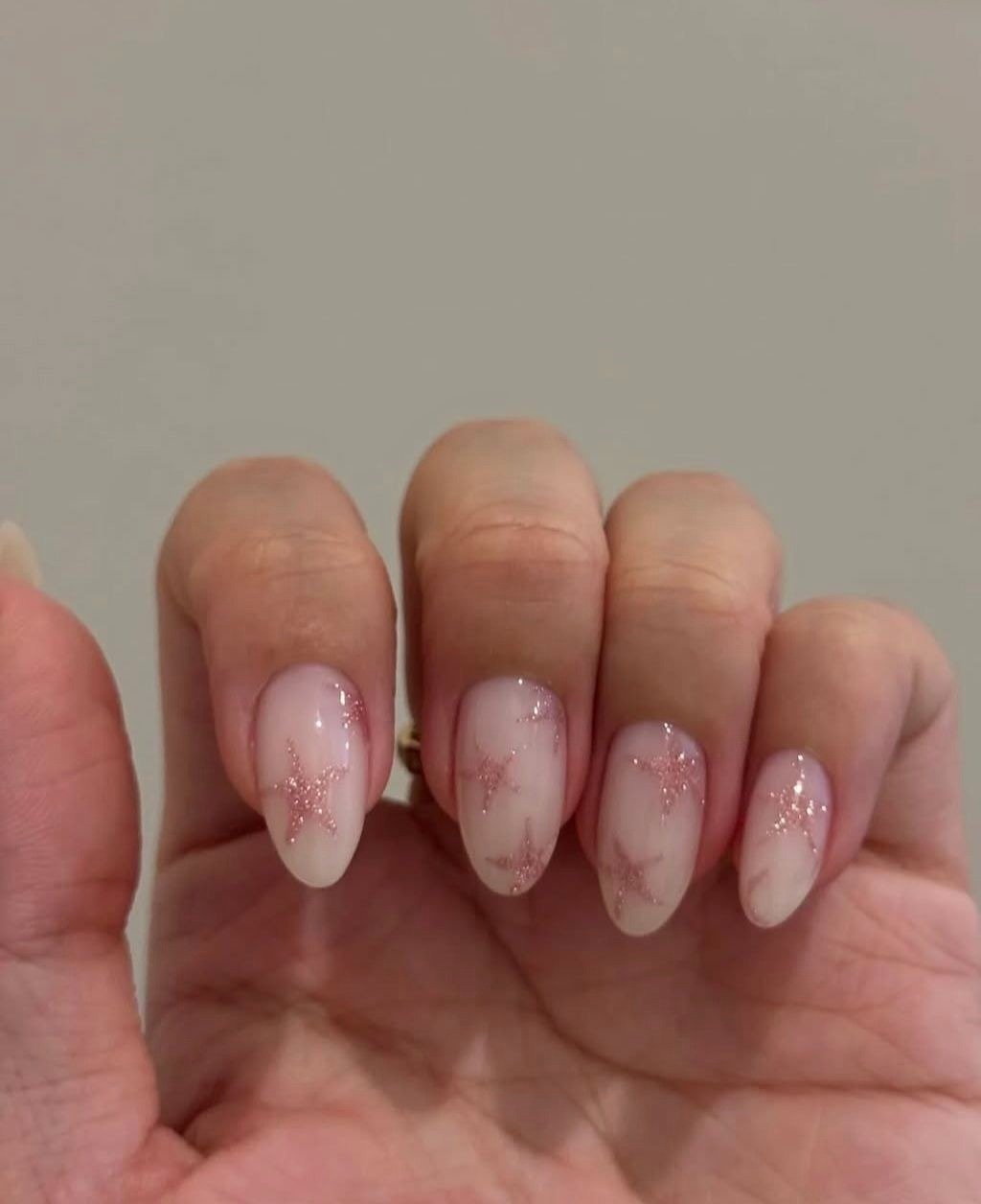 Unhas milky nails em tom leitoso com estrelas delicadas em glitter rosa, formato oval e acabamento brilhante, estilo suave e