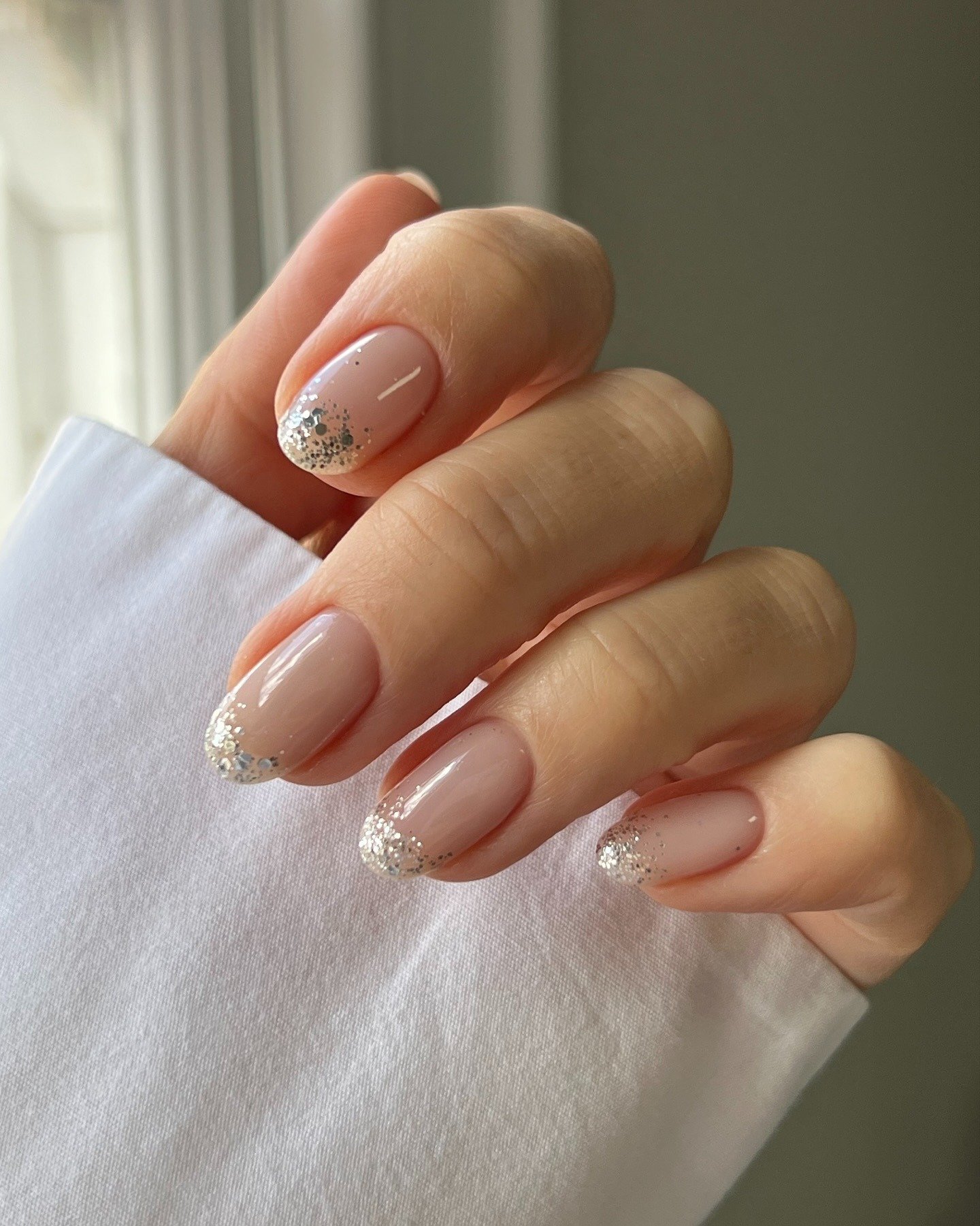 Unhas milky nails em tom nude leitoso com pontas delicadas de glitter prateado, mão repousa suavemente em tecido branco,