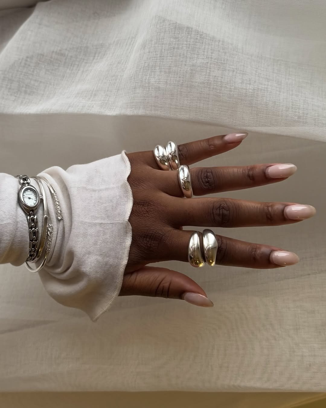 Mão com milky nails alongadas em tom leitoso, adornada por anéis largos prateados e pulseiras delicadas, sobre tecido branco