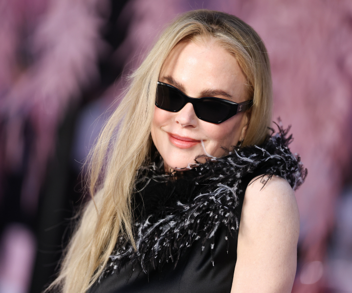 Atriz e produtora usa vestido preto com gola de penas texturizadas, óculos escuros e sorriso sereno em fundo desfocado rosa.