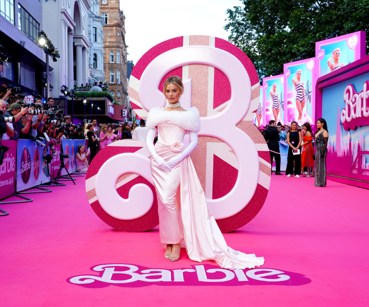 Atriz posa com vestido rosa acetinado, mangas longas e gola de plumas brancas, em tapete rosa vibrante da estreia de Barbie.