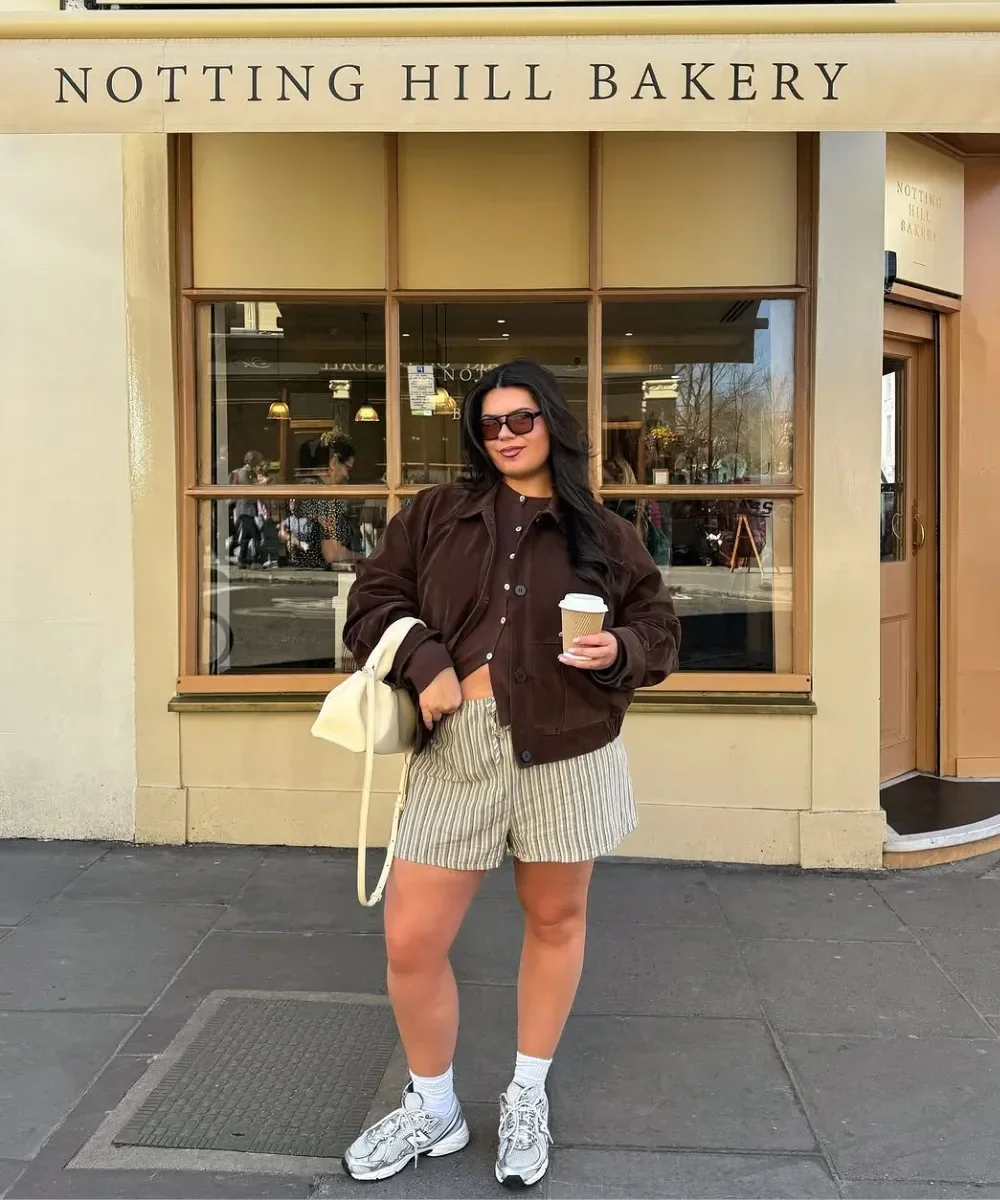 Modelo posa confiante em frente à padaria com tênis retrô prateado, shorts listrados, jaqueta marrom e bolsa creme, clima