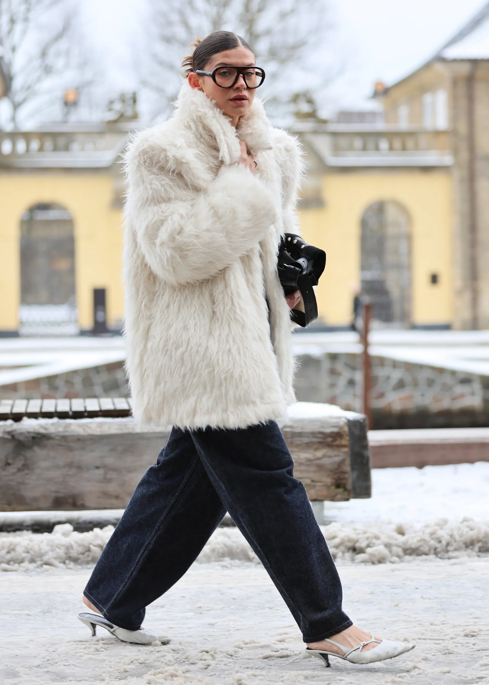 Modelo caminha confiante com óculos máscara oversized, casaco branco peludo, jeans escuros e mule branca em cenário urbano.