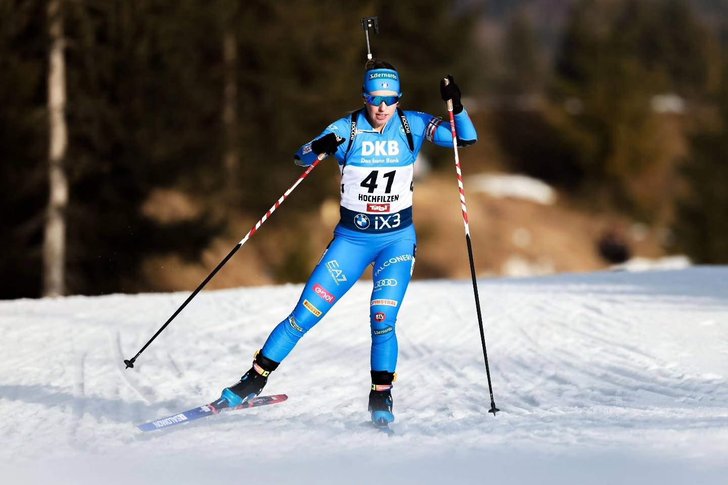 Atleta nas Olimpíadas de Inverno 2026 veste macacão azul justo com logos, esqui e bastões em ação dinâmica na neve