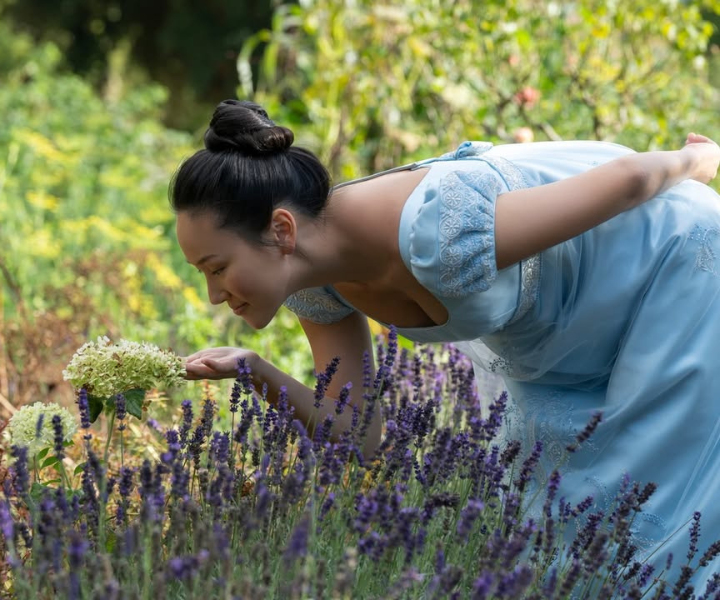 Modelo inclina-se para cheirar flores em vestido azul claro com bordados delicados, evocando perfumes florais e doces.