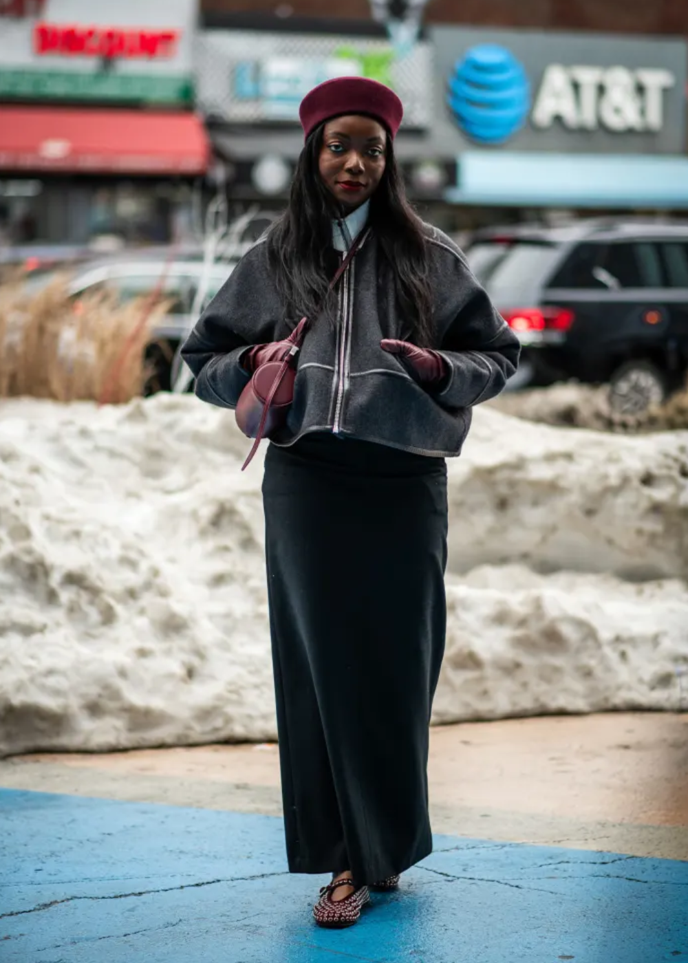 Modelo posa com pillbox hat vinho em tecido aveludado, jaqueta cinza oversized, saia longa preta e acessórios coordenados.