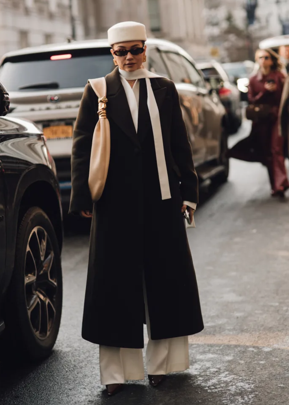 Modelo posa na rua com pillbox hat branco em tecido, sobretudo preto alongado, calça branca e bolsa bege.