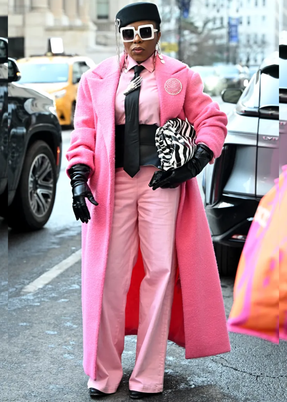 Modelo posa com pillbox hat preto adornado por pingentes prateados, casaco rosa oversized em lã, calça rosa e camisa rosa.