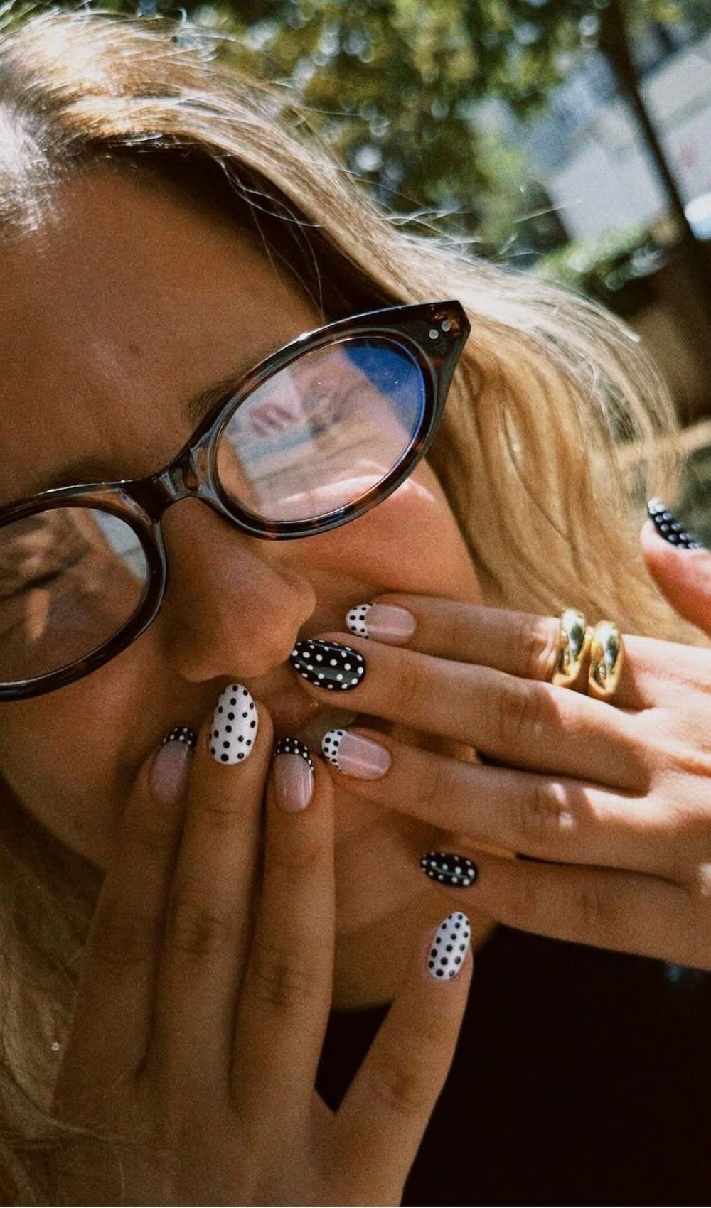 Unha de bolinhas em preto e branco com base nude e francesa, combinada a anéis dourados e óculos de armação tartaruga, em