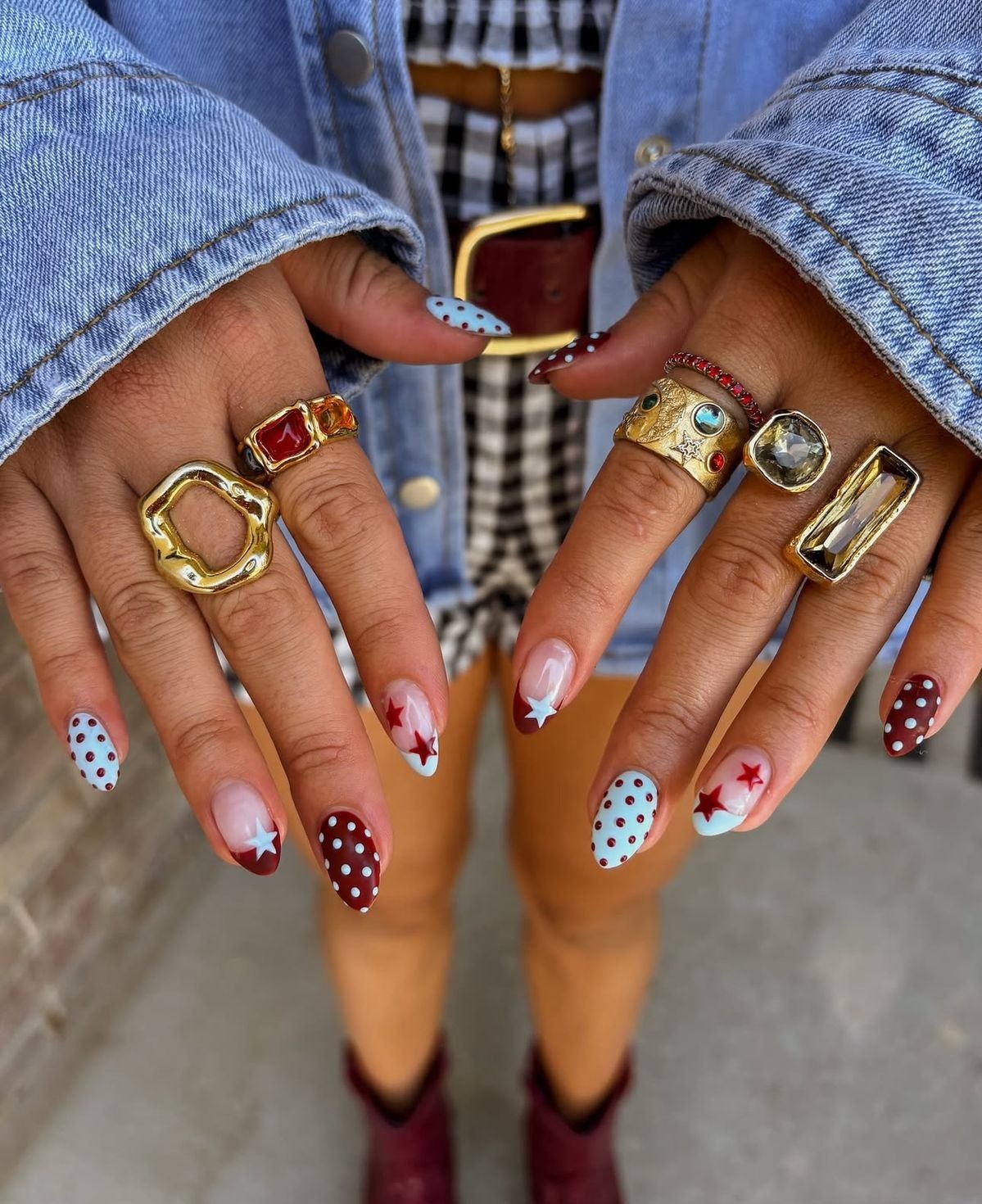 Modelo exibe unha de bolinhas em azul claro e vinho, com estrelas vermelhas, anéis dourados robustos e jaqueta jeans casual.