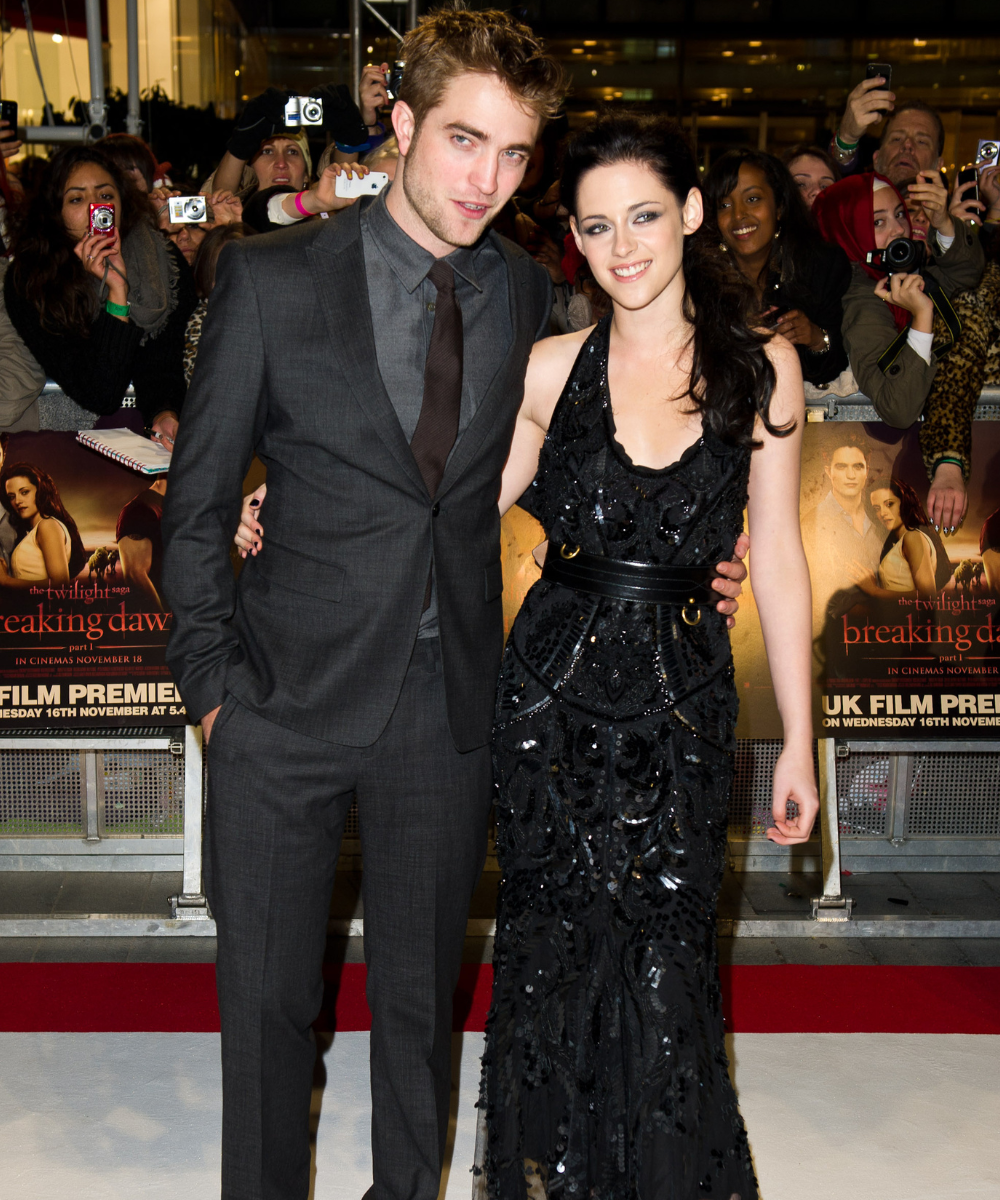 Casal posa abraçado no tapete vermelho com terno cinza de corte reto e vestido preto longo bordado em estilo PRomance, clima