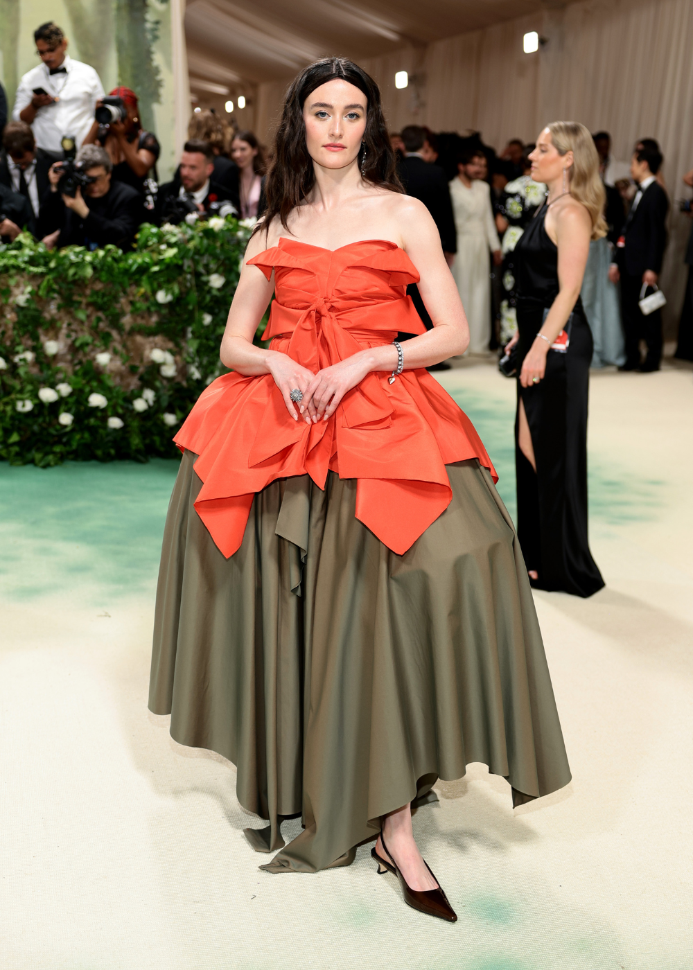 Sarah Pidgeon posa com vestido volumoso de tecido estruturado, corpete laranja com babados e saia ampla verde-oliva.