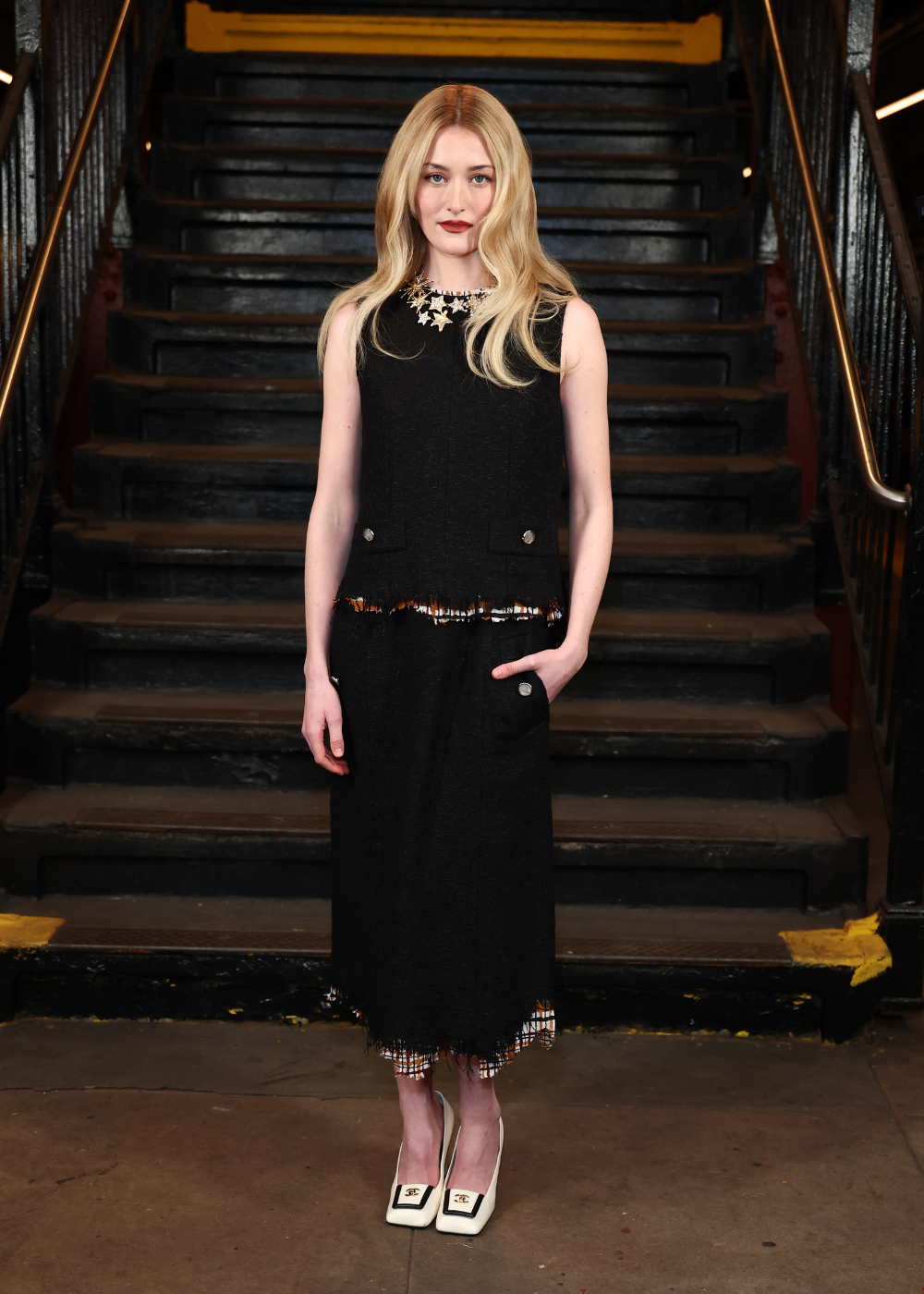 Sarah Pidgeon posa em vestido preto de tweed com detalhes desfiados e botões prateados, combinando scarpins brancos e colar.