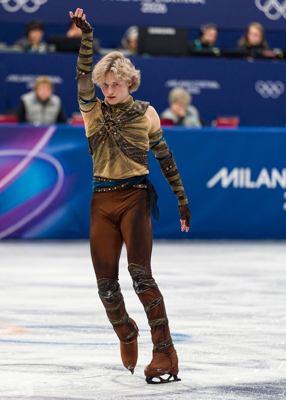Patinador masculino em pose dramática na patinação artística, vestindo traje justo marrom e dourado com detalhes brilhantes.