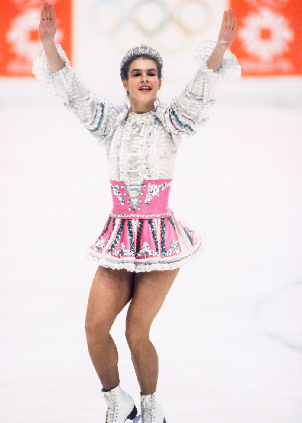 Patinadora artística celebra com postura ereta e braços erguidos, vestindo figurino branco e rosa com detalhes brilhantes.