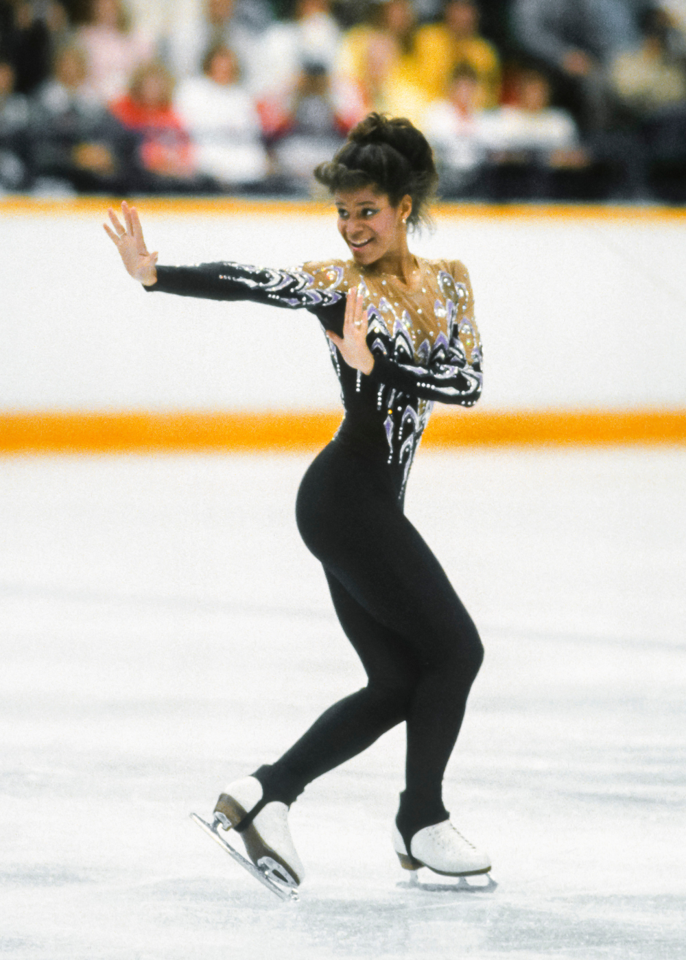 Patinadora artística executa pose elegante com macacão preto justo e detalhes brilhantes em tons neutros.