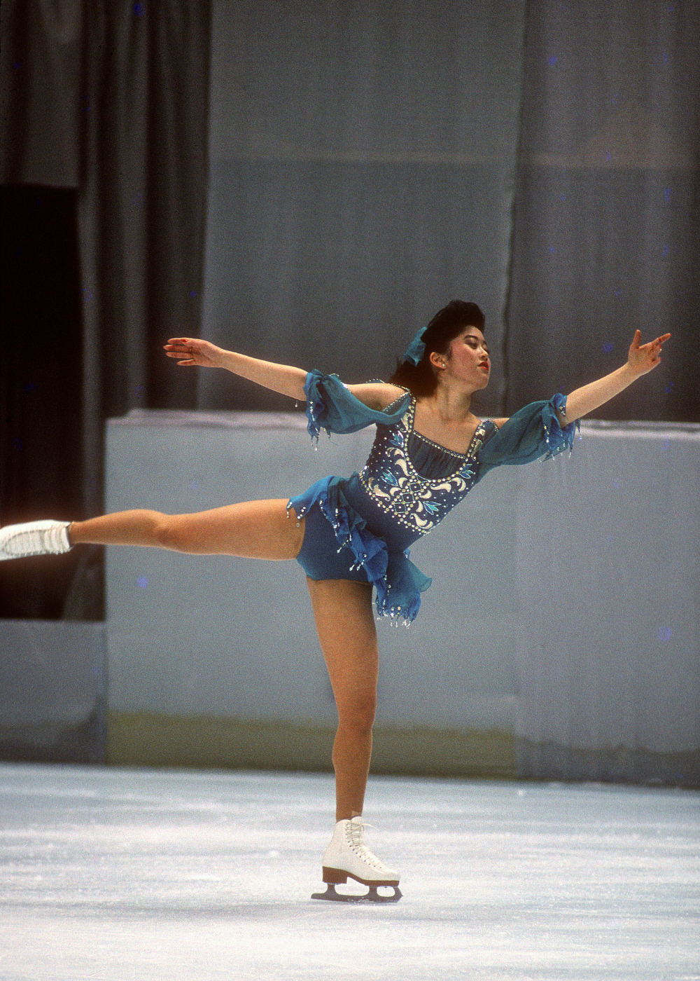 Patinadora artística executa pose elegante com vestido azul bordado, mangas fluídas e saia curta.