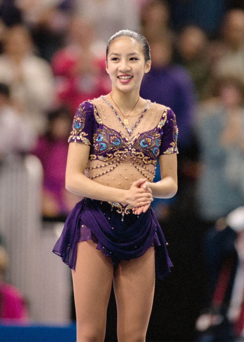 Patinadora artística sorri confiante com vestido roxo bordado em tule nude e saia fluida, destacando elegância e delicadeza.