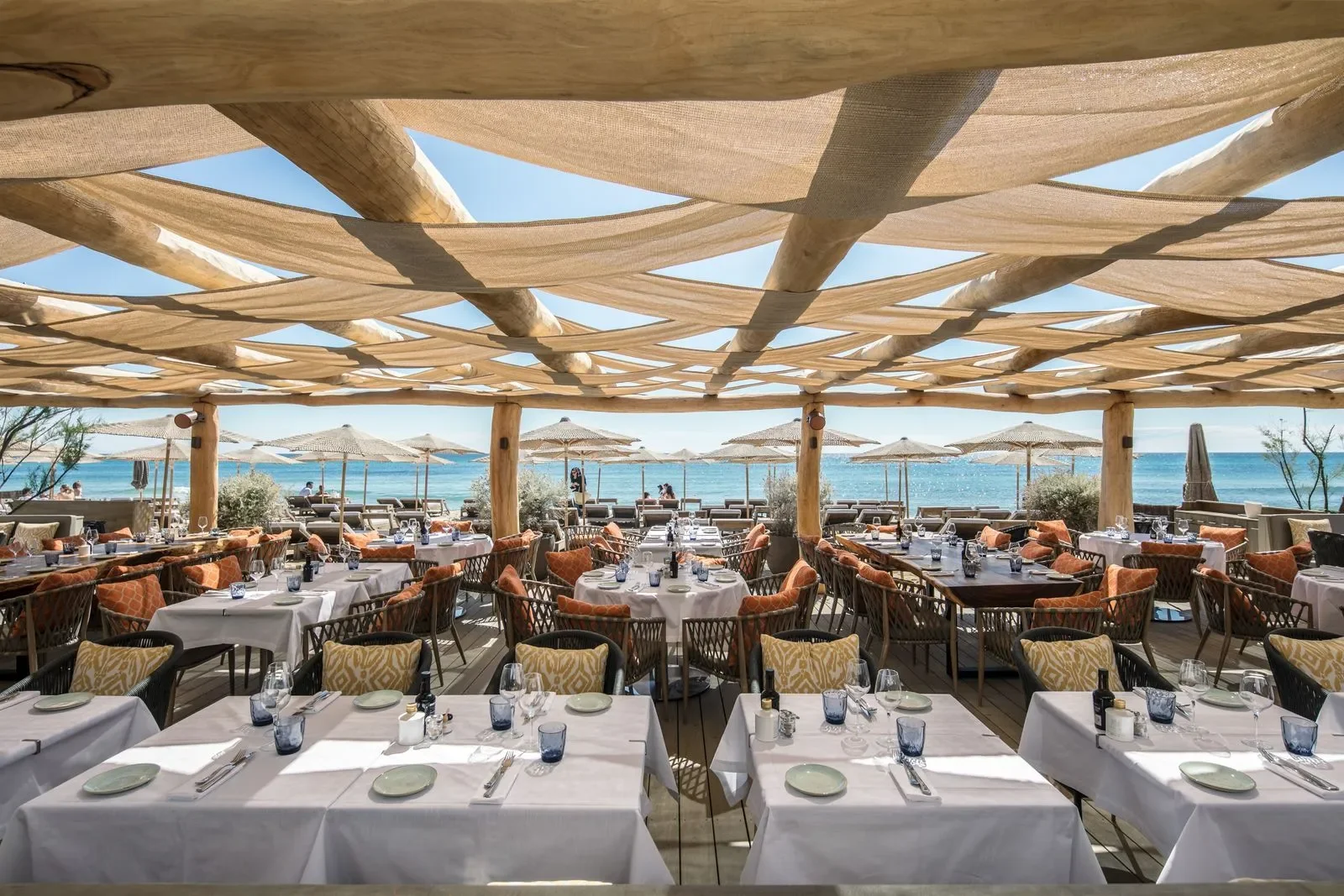 Restaurante à beira-mar em Saint-Tropez com mesas brancas, cadeiras de vime com almofadas laranja e bege, sob cobertura