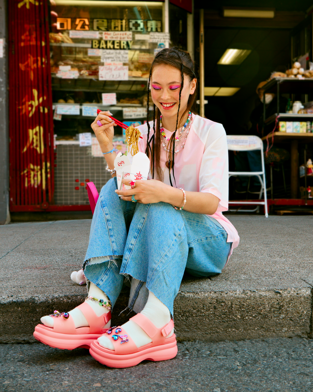 Modelo senta na calçada com sandália confortável e fashionista rosa vibrante, combinada com meia branca, jeans de barra