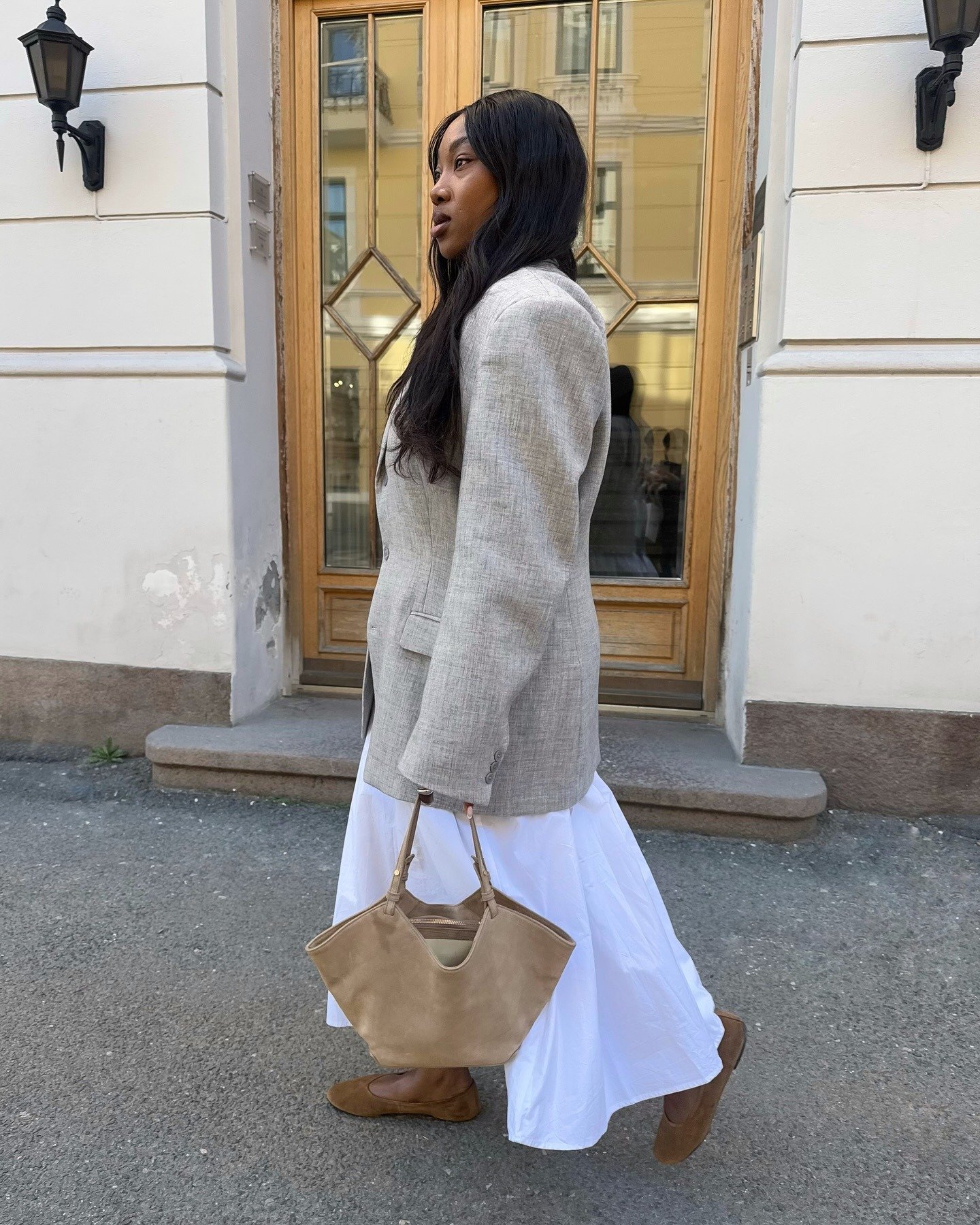 Modelo caminha confiante com blazer cinza oversized, saia branca fluida, bolsa bege e sapatilha tendência de 2026 em camurça