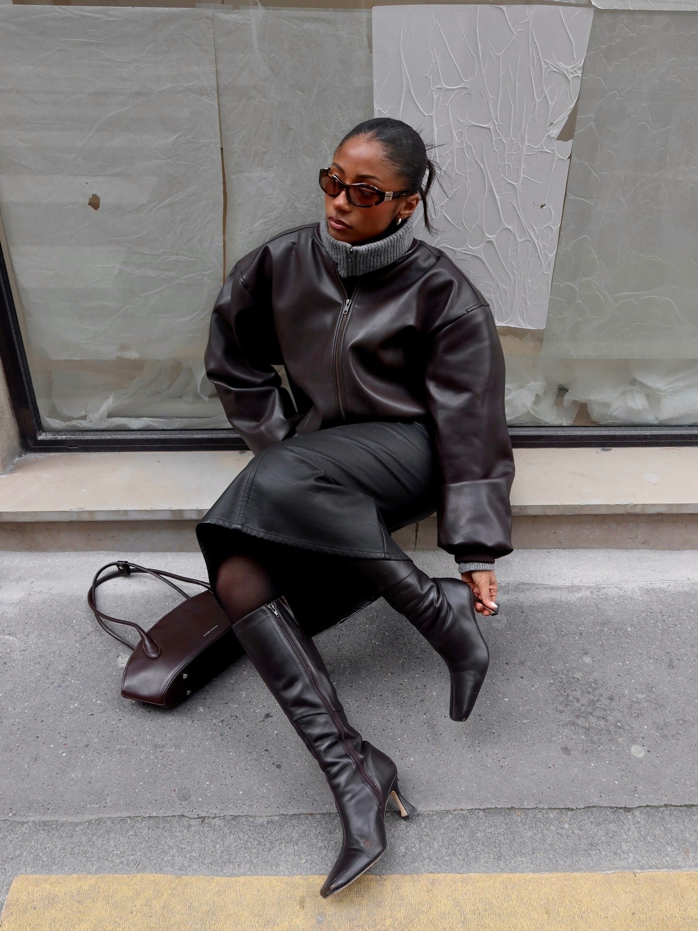 Modelo senta com postura confiante usando jaqueta oversized de couro marrom, saia midi e bota de cano alto