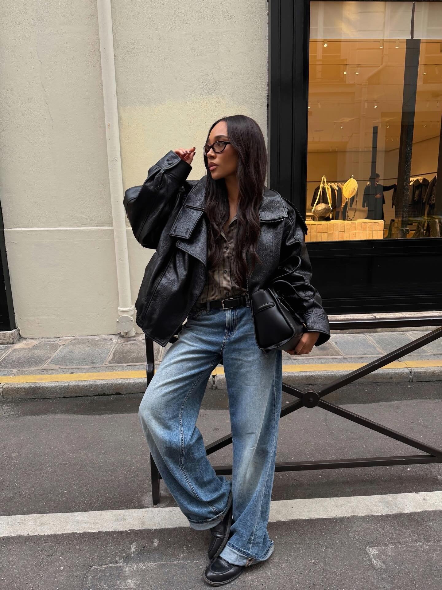 Modelo posa com jaqueta oversized de couro preto, camisa xadrez, jeans largos e bolsa estruturada, definindo a tendência