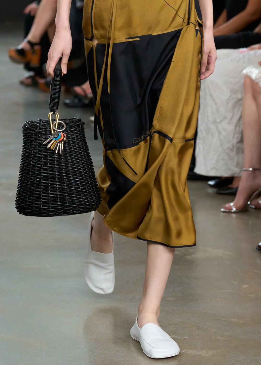 Modelo caminha com sapatos flat brancos de couro macio, saia midi fluida em cetim mostarda e preto.
