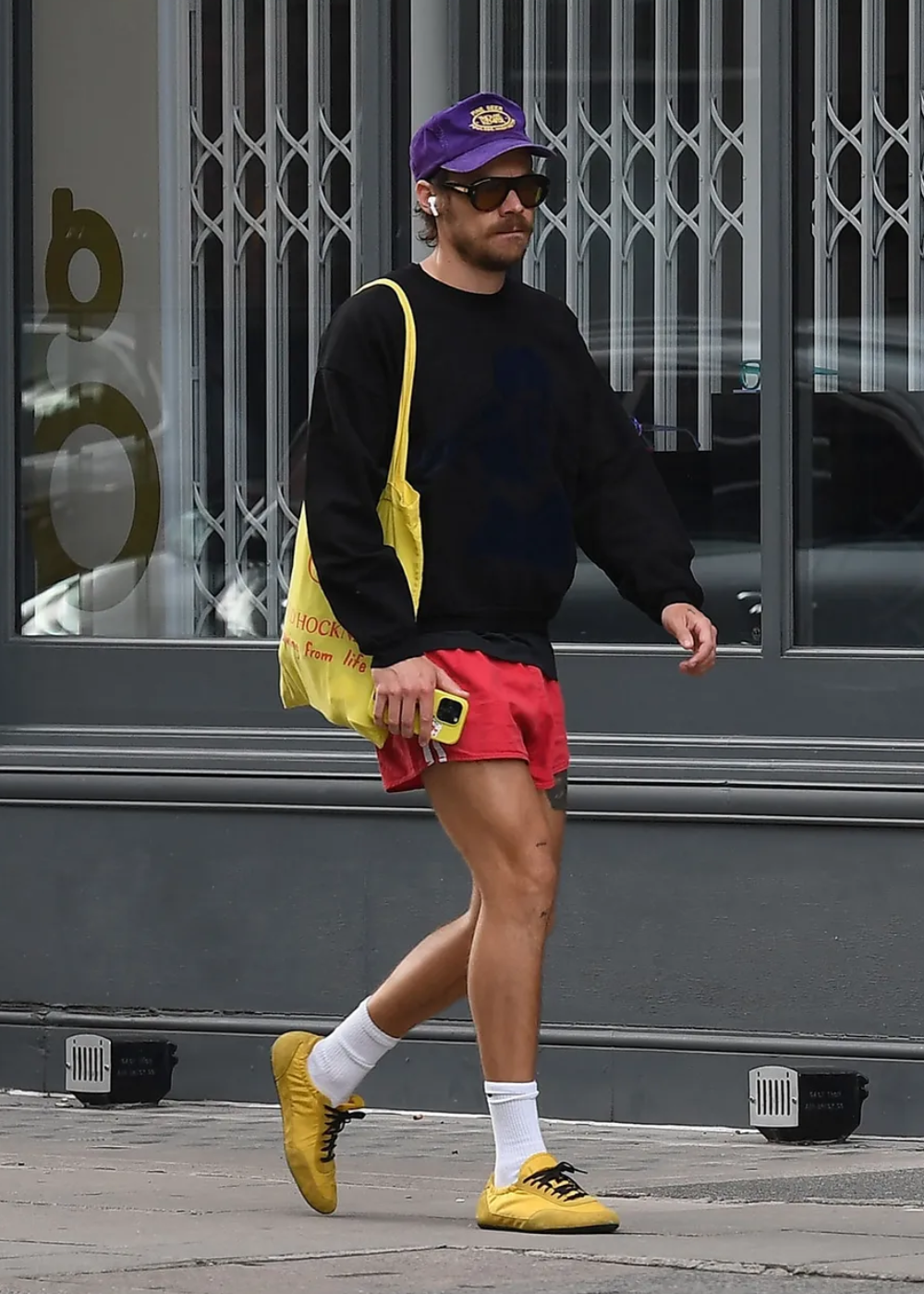 Homem caminha com sapatos flat amarelos de couro, shorts vermelhos, moletom preto e boné roxo em cenário urbano casual.