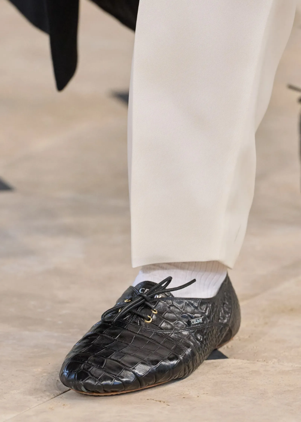 Sapatos flat em couro preto texturizado com amarração, combinados a calça branca e meia branca, evocando elegância discreta.