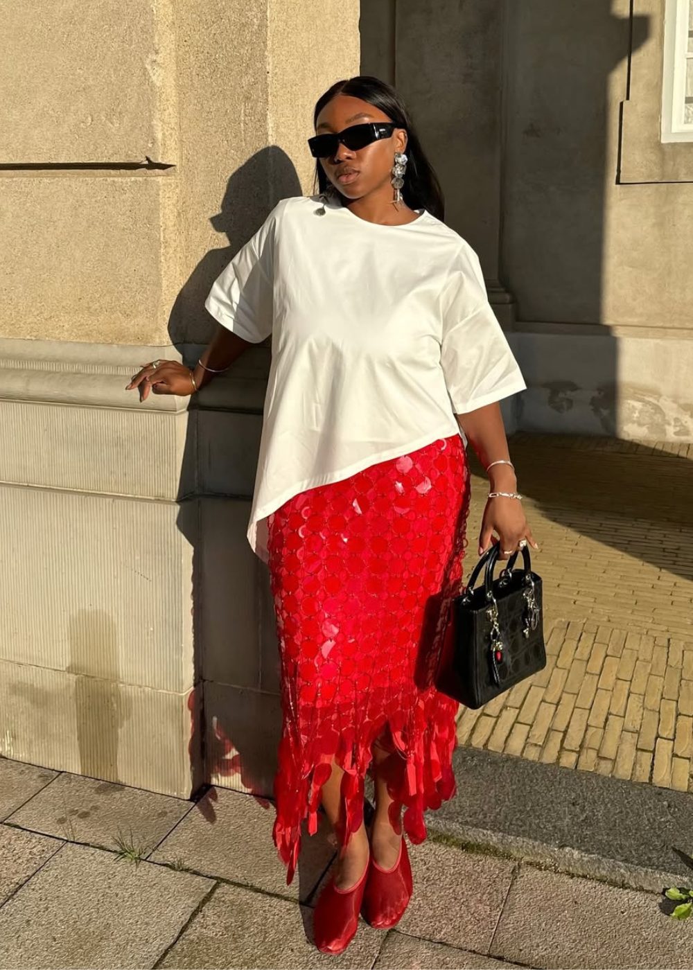 Modelo posa com blusa branca oversized, saia midi vermelha texturizada e sapatos flat vermelhos de tecido acetinado.